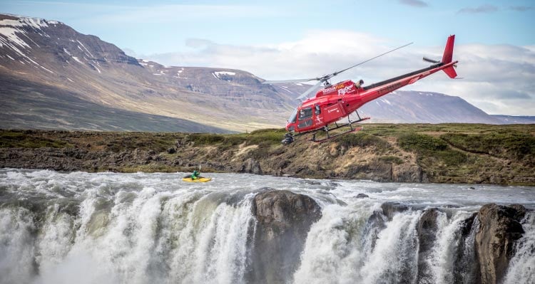A helicopter flies above a kayaker approaching a waterfall descent.