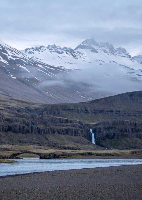 A waterfall descends towards a wide river below snow-covered mountains.