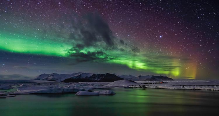 Green northern lights above snowy mountains and water.