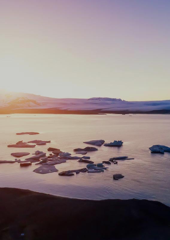 Large broken pieces of ice float in a cove as the sun sits behind a snowy mountain.