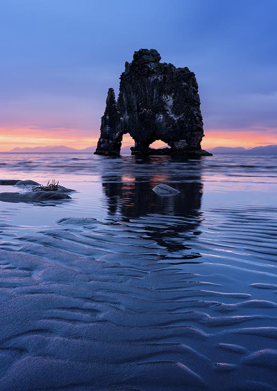 A tall rock formation in the water near a sandy beach