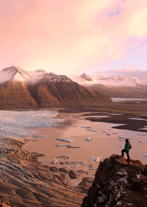 Göngumaður stendur fyrir ofan skriðjökul við sólsetur, með útsýni yfir hafið