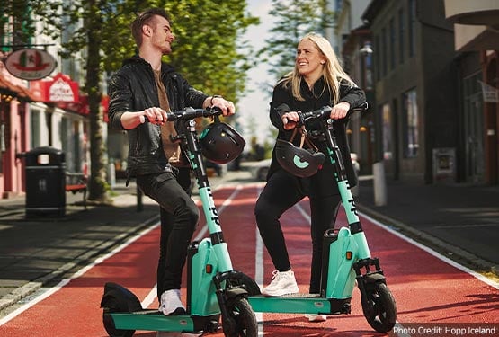 Two people riding e-Scooters on a leafy street.