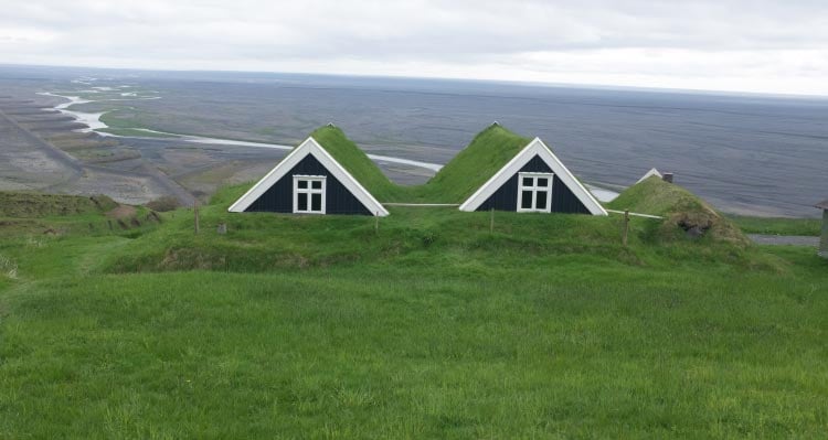 Two turf-house roofs peak outside of a green hill, overlooking a river.