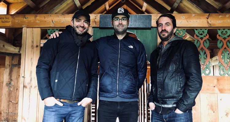 Thee men stand in a rustic wooden church with intricate decorations.