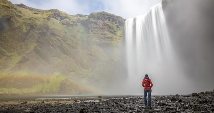 A person in a red jacket stands before a large waterfall.