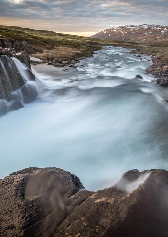 A river rushes over a small cliff into a wide green land.