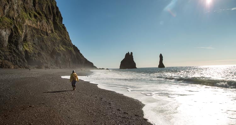 A person walks along a beach between the ocean and a cliffside.