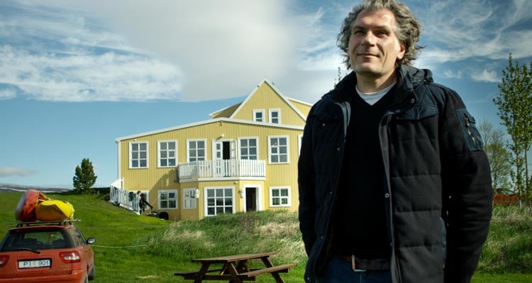 A man stands outside a yellow house under a blue and cloudy sky.