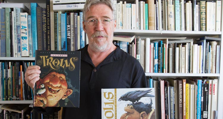 Brian Pilkington holds two of his Icelandic Trolls books in front of a bookshelf