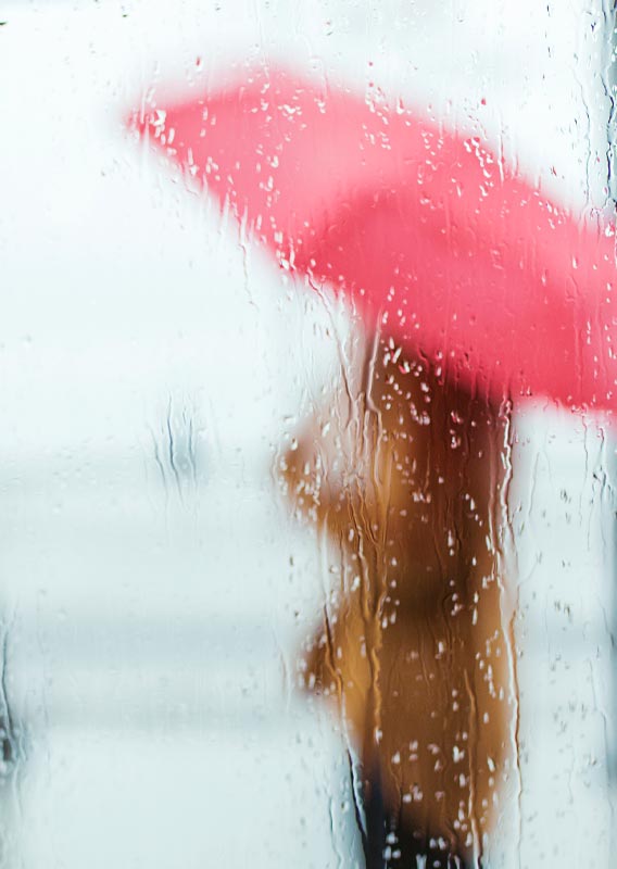 A person with a red umbrella stands behind a rain-covered window.