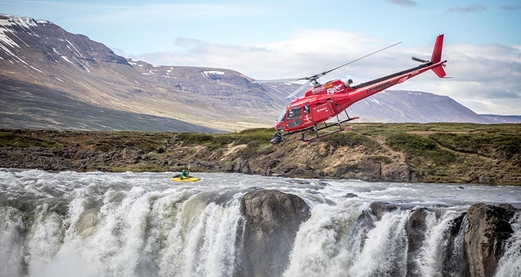 Kajak ræðari stendur fyrir ofan foss. Rauð þyrla flýgur að.