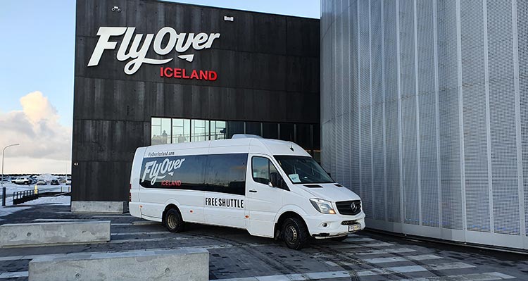A minibus parked in front of the silver and black FlyOver Iceland building.