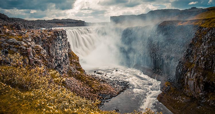 The waterfall Dettifoss. A tall and wide waterfall crashes into mist below mossy and rocky cliffsides.