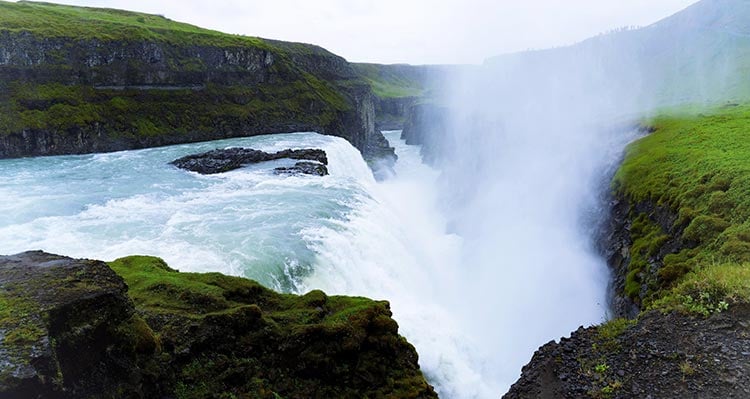 The waterfall Gullfoss. A waterfall crashes into a misty pool below grassy hillsides.