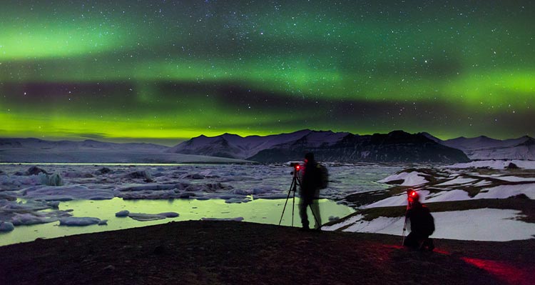 Two people watch green northern lights above an icy sea.