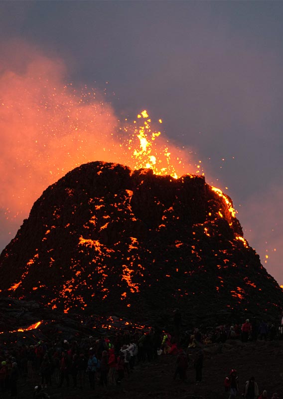 Volcano spews lava, people a large group of people watch form afar.
