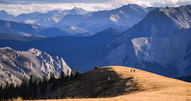 Two horseback riders look over a grassy cliff to a mountain range.