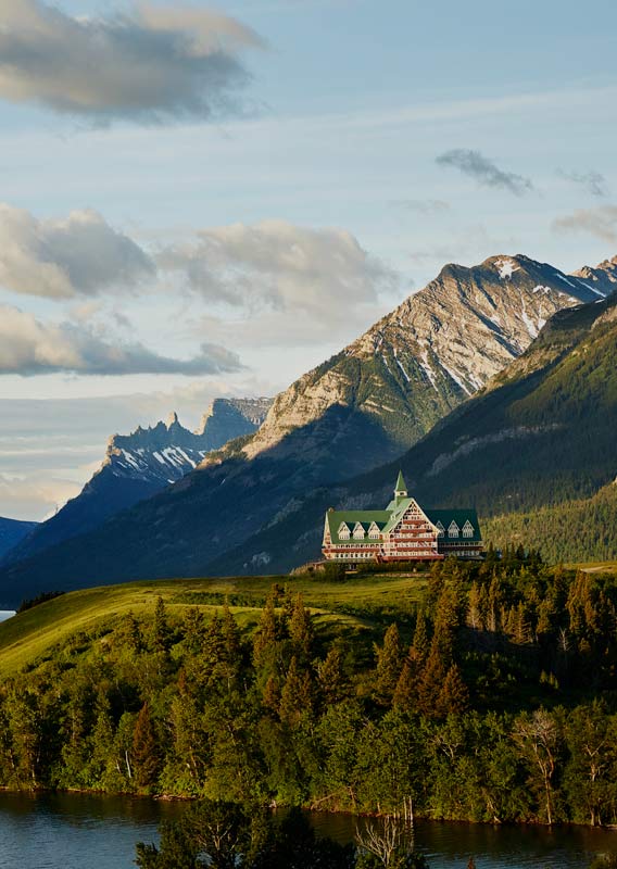 A large wooden lodge building atop a grassy hill above a large blue lake below tall mountains.