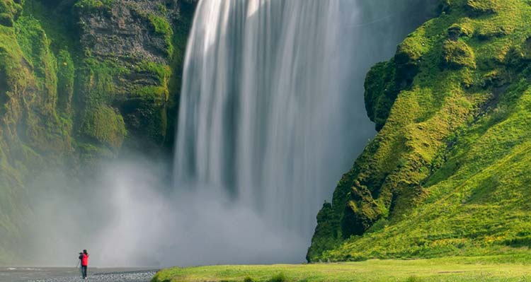 A hiker in a red jacket stands below a giant waterfall.